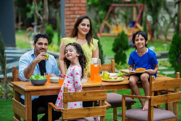 indian young family of four eating meal on dining table in the garden
