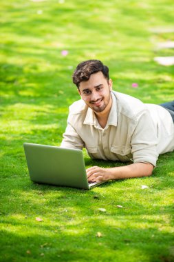 Genç, yakışıklı Hintli Asyalı adam çimlerin üzerinde yatarken parkta dizüstü bilgisayar kullanıyor.
