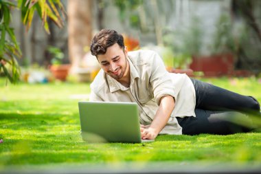 Genç, yakışıklı Hintli Asyalı adam çimlerin üzerinde yatarken parkta dizüstü bilgisayar kullanıyor.