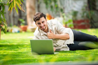 Genç, yakışıklı Hintli Asyalı adam çimlerin üzerinde yatarken parkta dizüstü bilgisayar kullanıyor.