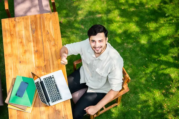 Overhead view of handsome indian man using laptop in the garden at table looking up into camera