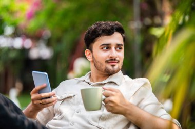 Hintli genç adam dışarıda oturuyor ve akıllı telefon kullanıyor.