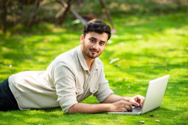 Genç, yakışıklı Hintli Asyalı adam çimlerin üzerinde yatarken parkta dizüstü bilgisayar kullanıyor.
