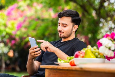 Hintli genç adam dışarıda oturuyor ve akıllı telefon kullanıyor.