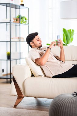 Genç Hintli yakışıklı adam oturma odasında kanepede otururken akıllı telefon kullanıyor.