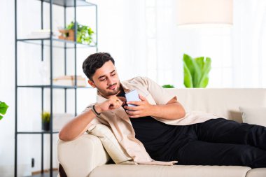 Genç Hintli yakışıklı adam oturma odasında kanepede otururken akıllı telefon kullanıyor.