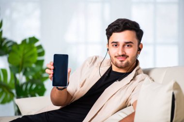 Genç Hintli yakışıklı adam oturma odasında kanepede otururken akıllı telefon kullanıyor.