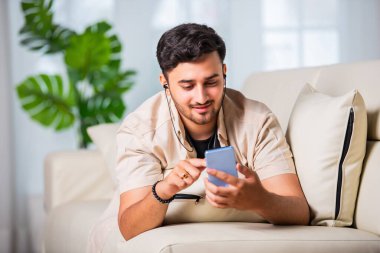 Genç Hintli yakışıklı adam oturma odasında kanepede otururken akıllı telefon kullanıyor.