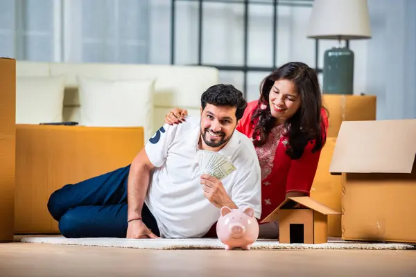 Indian couple saving money in piggybank while sitting in their new home