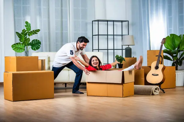 Energetic happy Indian wife sitting in cardboard container, having fun while moving day and husband pushing her