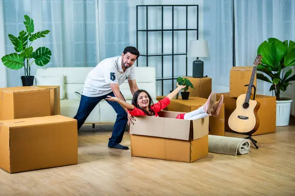 Energetic happy Indian wife sitting in cardboard container, having fun while moving day and husband pushing her