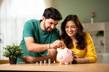 Hintli genç çift ve tasarruf konsepti madeni para ve domuz kumbarası kullanıyorlar.