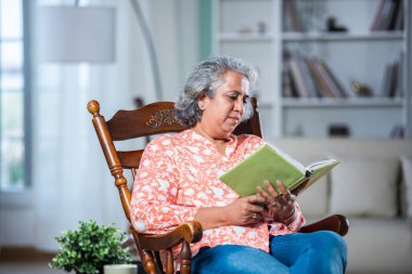 Hintli orta yaşlı gri saçlı kadın oturma odasında sallanan sandalyede otururken kitap okuyor.