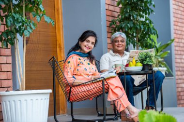 Yaşlı Hintli çift gazete ve kitap okurken müzik dinliyor ve dışarıda galeride kahvaltı yapıyor.