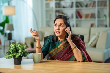 60 'lı yaşlarda Hintli bir kadın evde akıllı telefon kullanıyor, kahvaltı masasında oturuyor.