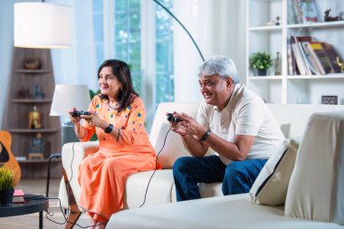 Hintli yaşlı çift evdeki kanepede otururken video oyunu oynayarak eğleniyor.