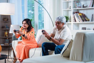 Hintli yaşlı çift evdeki kanepede otururken video oyunu oynayarak eğleniyor.