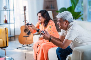 Hintli yaşlı çift evdeki kanepede otururken video oyunu oynayarak eğleniyor.