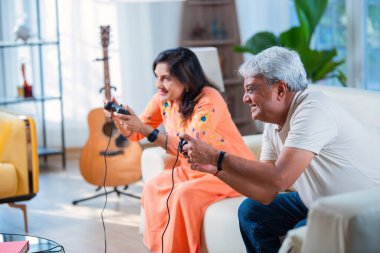 Hintli yaşlı çift evdeki kanepede otururken video oyunu oynayarak eğleniyor.