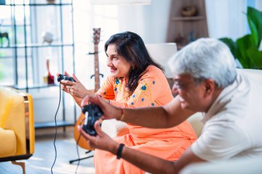Hintli yaşlı çift evdeki kanepede otururken video oyunu oynayarak eğleniyor.