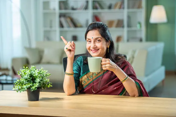 Retired Indian asian senior woman in saree having coffee or tea at home on breakfast table
