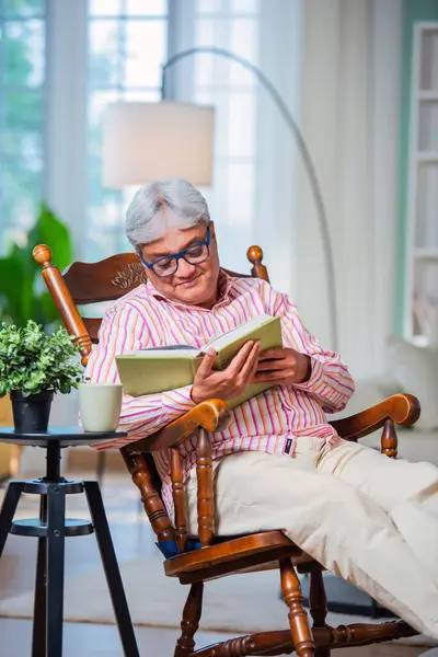 Indian asian Old man reading book or novel while sitting on rocking chair at home