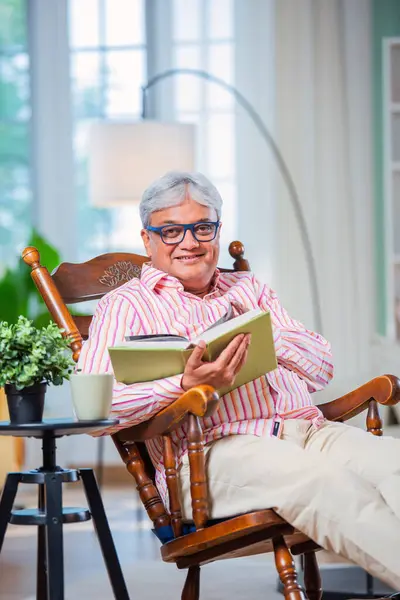 Indian asian Old man reading book or novel while sitting on rocking chair at home