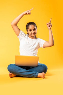 Hintli küçük kız dizüstü bilgisayar kullanıyor sarı arka planda tek başına oturuyor.