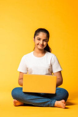 Hintli küçük kız dizüstü bilgisayar kullanıyor sarı arka planda tek başına oturuyor.