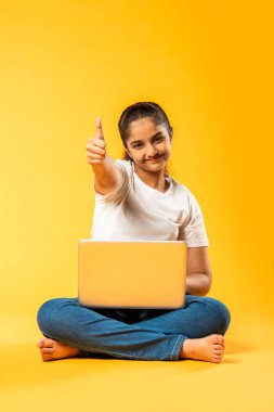 Hintli küçük kız dizüstü bilgisayar kullanıyor sarı arka planda tek başına oturuyor.