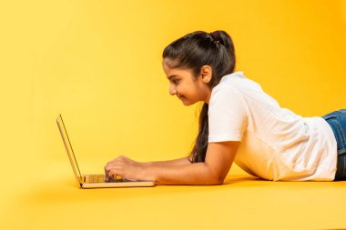 Sarı zeminde yatarken dizüstü bilgisayar kullanan Hintli küçük kız tecrit edilmiş.