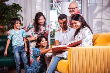 Hintli Asyalı çok nesilli aile birlikte kanepede otururken fotoğraf izliyor ya da kitap okuyor.