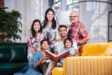 Hintli Asyalı çok nesilli aile birlikte kanepede otururken fotoğraf izliyor ya da kitap okuyor.