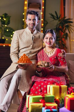 Hintli evli genç çift diwali festivalini kutluyor puja thali ve şeker tutuyorlar kanepede otururken.