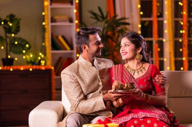 Hintli evli genç çift diwali festivalini kutluyor puja thali ve şeker tutuyorlar kanepede otururken.