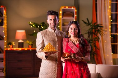 Hintli evli genç çift diwali festivalini kutluyor puja thali ve şeker tutuyorlar kanepede otururken.