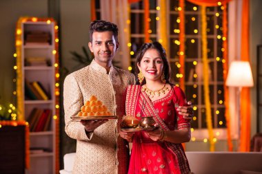 Hintli evli genç çift diwali festivalini kutluyor puja thali ve şeker tutuyorlar kanepede otururken.