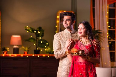 Hintli evli genç çift diwali festivalini kutluyor puja thali ve şeker tutuyorlar kanepede otururken.