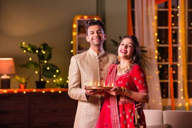 Hintli evli genç çift diwali festivalini kutluyor puja thali ve şeker tutuyorlar kanepede otururken.