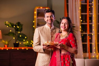 Hintli evli genç çift diwali festivalini kutluyor puja thali ve şeker tutuyorlar kanepede otururken.