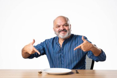 Masada otururken elinde boş seramik tabak tutan Hintli emekli yaşlı adam.