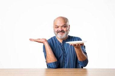 Masada otururken elinde boş seramik tabak tutan Hintli emekli yaşlı adam.