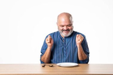 Masada otururken elinde boş seramik tabak tutan Hintli emekli yaşlı adam.