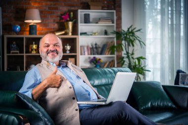 Hintli yakışıklı emekli yaşlı adam kanepede otururken akıllı telefon ya da laptop kullanıyor.