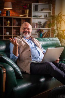 Hintli yakışıklı emekli yaşlı adam kanepede otururken akıllı telefon ya da laptop kullanıyor.