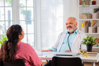 Hintli yaşlı yaşlı yaşlı doktor, klitonik giyinmiş kadın hastayla durumu tartışıyor.