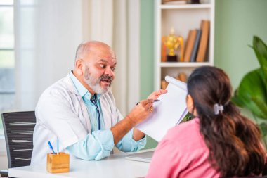 Hintli yaşlı yaşlı yaşlı doktor, klitonik giyinmiş kadın hastayla durumu tartışıyor.