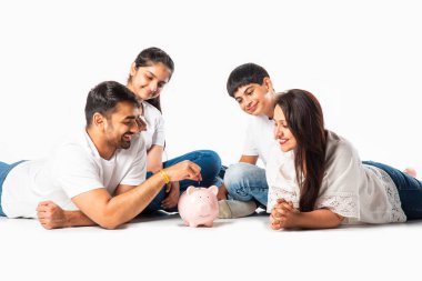 Aile Ekonomisi. Mutlu Asyalı anne, baba ve iki çocuk domuzcuk kumbarasıyla yerde yatıp kameraya gülümsüyor.