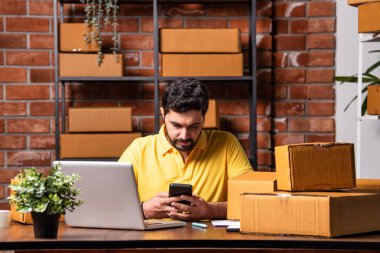 Hintli Asyalı Adam Dizüstü bilgisayarlı ve akıllı telefonlu online siparişleri yönetiyor, Depo 'dan gönderilmeye hazır.