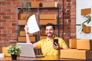 Hintli Asyalı Adam Dizüstü bilgisayarlı ve akıllı telefonlu online siparişleri yönetiyor, Depo 'dan gönderilmeye hazır.